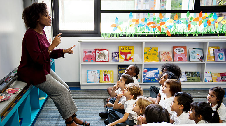 Primary school teacher explains at the front of a class while school children sit cross-legged watching and listening